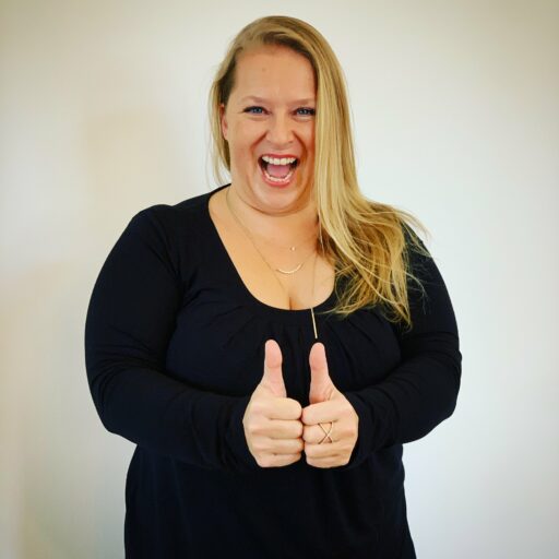 Frau mit langen blonden Haaren strahlt in die Kamera und hat beide Daumen vor sich in der Höhe